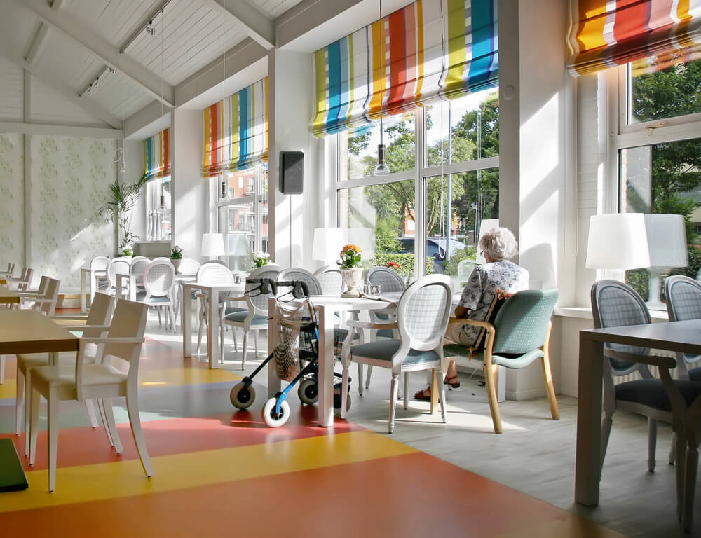 Elderly lady sitting in a large room full of tables and chairs with a walking aid next to her.