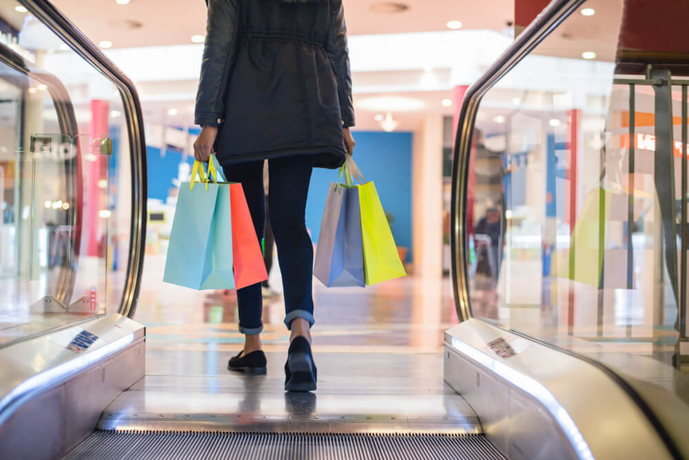 Person about to go down an escalator in a shopping centre carrying shopping bags.